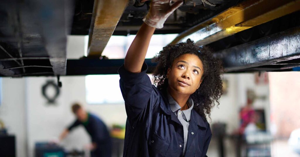 A young female adult mechanic is standing under a car in a garage. She is standing proudly and looking to camera